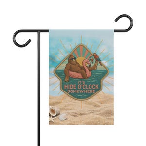 Könnte beinhalten: Eine Gartenflagge mit einem humorvollen Design. Sie zeigt einen Cartoon-Bigfoot mit Sonnenbrille, der auf einem Flamingo-Schwimmer liegt und ein Getränk hält. Die Flagge trägt den Text "IT'S HIDE O'CLOCK SOMEWHERE" vor einem Strandhintergrund.
