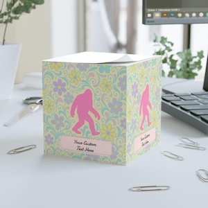 Puede incluir: Un bloc de notas cuadrado con un estampado floral en colores pastel, con siluetas rosas de Bigfoot. El bloc de notas tiene una etiqueta rectangular rosa claro con el texto "Your Custom Text Here".