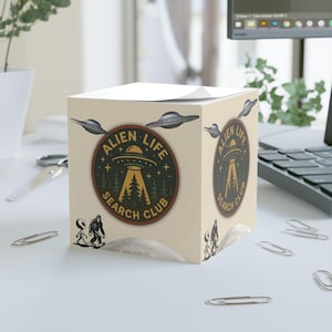 May include: A beige cube-shaped notepad with an "Alien Life Search Club" design. The design features a UFO, trees, and the text "Alien Life Search Club". The cube also has illustrations of UFOs and aliens.