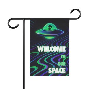 May include: A rectangular garden flag hanging on a black metal stand. The flag features a glowing green UFO with an alien silhouette inside, set against a dark background with wavy purple and green lines and the text WELCOME TO OUR SPACE.