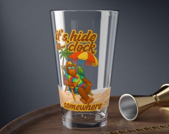 May include: A clear pint glass with a beach-themed graphic. The design features a cartoon bear in a tie-dye shirt relaxing under an umbrella, with the text "it's hide o'clock somewhere". A gold bar spoon and jigger are on the wooden surface.