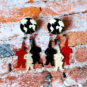 May include: A pair of dangle earrings with a black and white cow print design. Each earring has three charms: a red, a white, and a black figure.