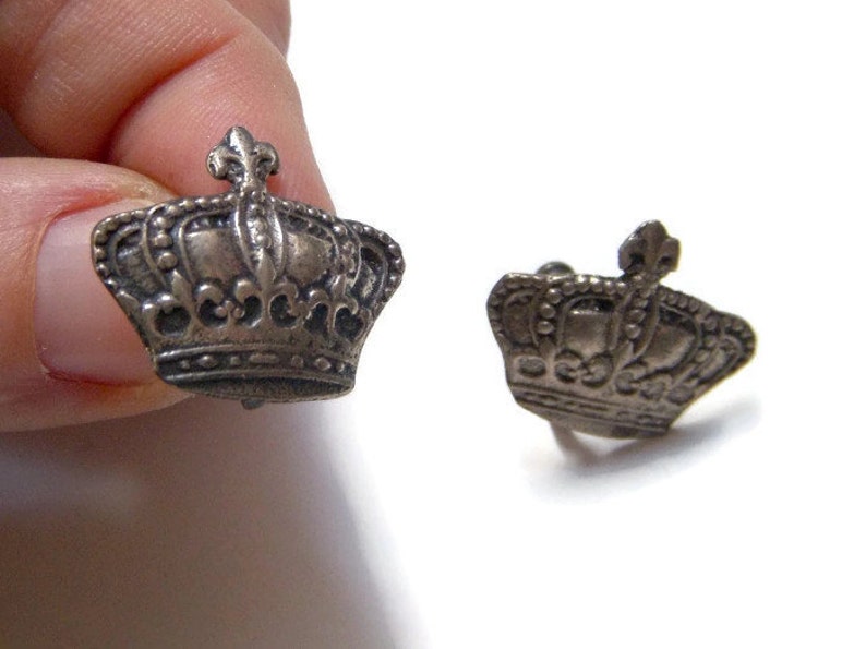 K&ouml;nnte beinhalten: Ein Paar silberne Manschettenkn&ouml;pfe in Kronenform. Die Kronen sind mit komplizierten Designs verziert und haben eine leicht gealterte Oberfl&auml;che.