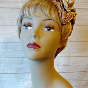 K&ouml;nnte beinhalten: Ein Mannequin-Kopf mit blonden Haaren, der ein braunes und wei&szlig;es Stoffband mit einer wei&szlig;en Rose und gr&uuml;nen Bl&auml;ttern tr&auml;gt.