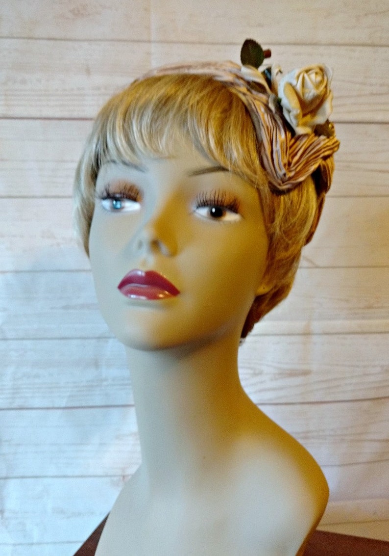 K&ouml;nnte beinhalten: Ein Mannequin-Kopf mit blonden Haaren, der ein braunes und wei&szlig;es Stoffband mit einer wei&szlig;en Rose und gr&uuml;nen Bl&auml;ttern tr&auml;gt.