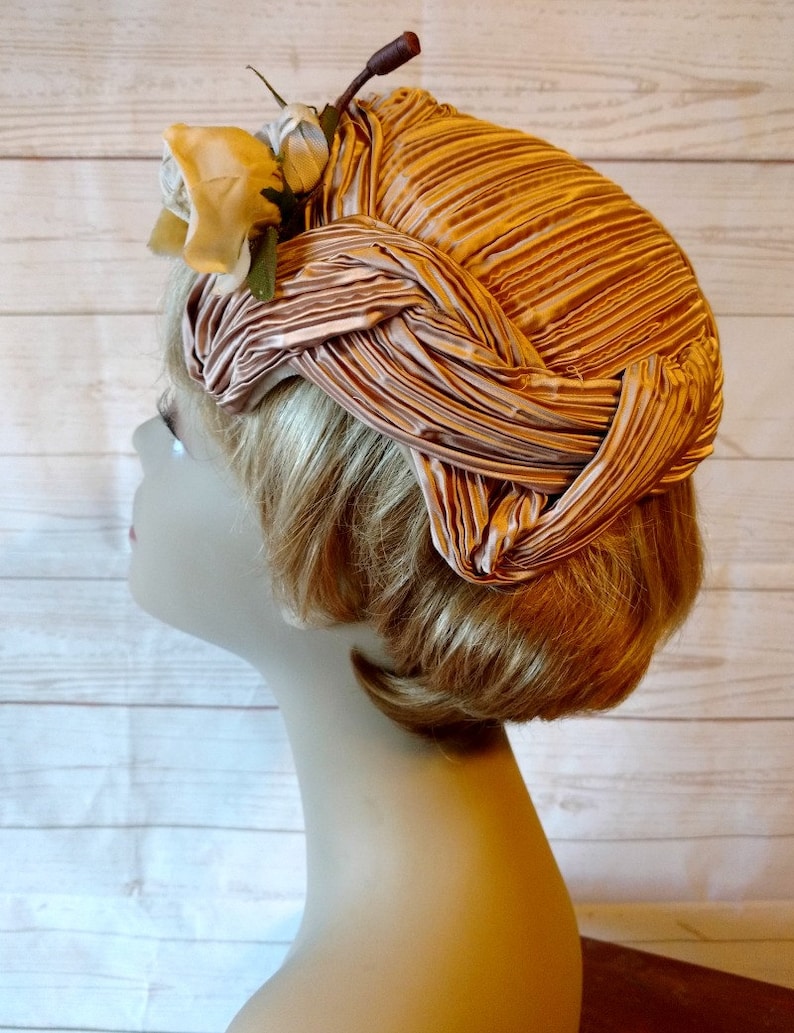 K&ouml;nnte beinhalten: Ein rosa gefalteter Hut aus Stoff mit einem wei&szlig;en Blumenakzent. Der Hut wird auf einem Mannequin-Kopf mit blonden Haaren getragen.
