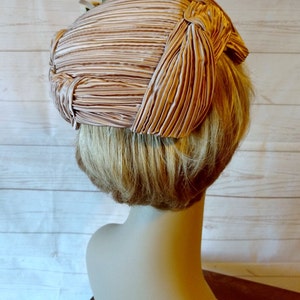 K&ouml;nnte beinhalten: Ein rosa gefalteter Hut mit einer Schleife oben. Der Hut sitzt auf einem Mannequin-Kopf mit blonden Haaren.