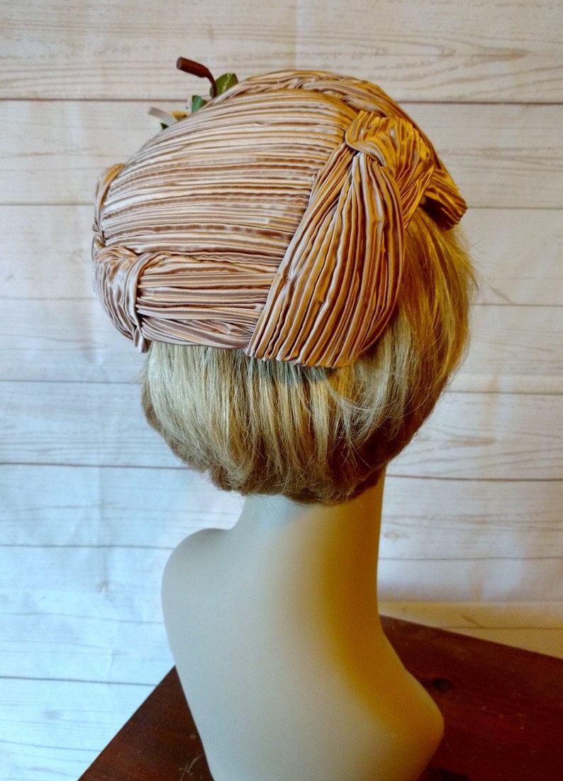 K&ouml;nnte beinhalten: Ein rosa gefalteter Hut mit einer Schleife oben. Der Hut sitzt auf einem Mannequin-Kopf mit blonden Haaren.