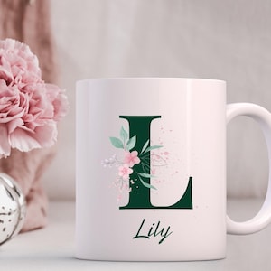 May include: White ceramic mug featuring a dark green letter "L" adorned with pink floral accents and green leaves. The name "Lily" is written in a script font below the letter. The mug has a white handle.