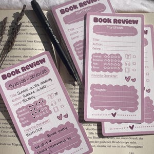 Könnte beinhalten: Eine Ansammlung von rosa-weißen Buchrezensionskarten und ein schwarzer Stift auf einem offenen Buch. Die Karten bieten Platz für Titel, Autor und Genre, sowie ein Bewertungssystem und Notizen. Eine Karte trägt den Titel "Sunrise on the Reaping".