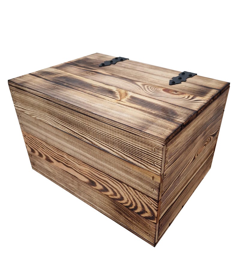 May include: A rustic wooden storage box with a hinged lid and black metal hardware. The box is constructed from light brown wood planks with a visible grain pattern and darker, burnt-looking edges. The box is rectangular and appears to be handmade.