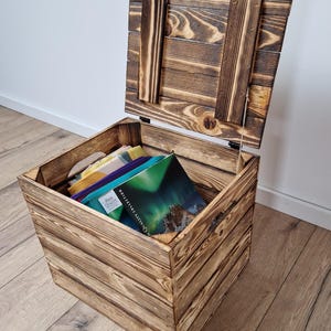 May include: A rustic wooden storage chest with a hinged lid, partially open. The chest is made of light brown wood with visible grain and a distressed finish. Inside, colorful items and a book are visible. The chest is on a wooden floor.
