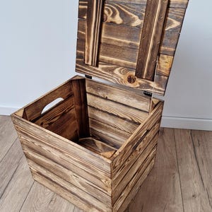 May include: A rustic wooden storage box with a hinged lid, crafted from light brown planks. The lid is slightly ajar, revealing the interior. The box features cut-out handles on the sides, and is set on a wooden floor.