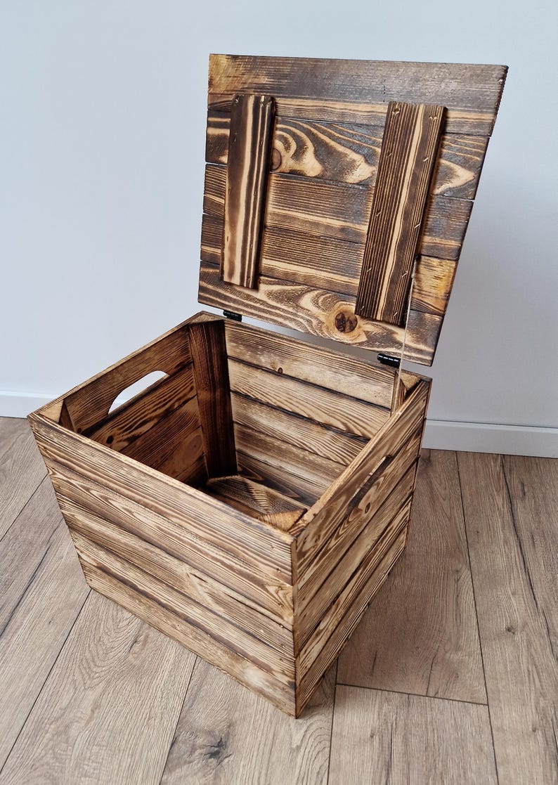 May include: A rustic wooden storage box with a hinged lid, crafted from light brown planks. The lid is slightly ajar, revealing the interior. The box features cut-out handles on the sides, and is set on a wooden floor.