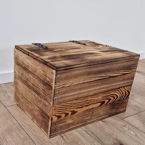 May include: A square wooden storage box with a hinged lid. The box is constructed from planks of wood with a visible grain pattern and a dark, burnt finish. The hinges are black metal.