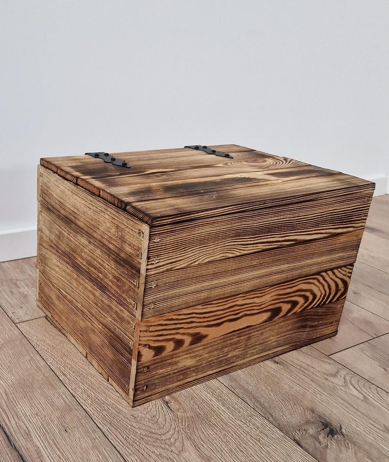 May include: A square wooden storage box with a hinged lid. The box is constructed from planks of wood with a visible grain pattern and a dark, burnt finish. The hinges are black metal.