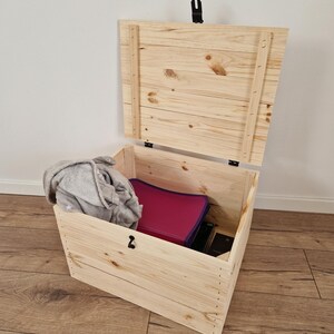 K&ouml;nnte beinhalten: Eine offene, unfertige Holzkiste mit Scharnierdeckel. Im Inneren sind eine graue Decke und ein magenta Ordner zu sehen. Die Kiste ist aus hellem Holz gefertigt und hat einen schwarzen Verschluss. Die Kiste steht auf einem Holzboden.