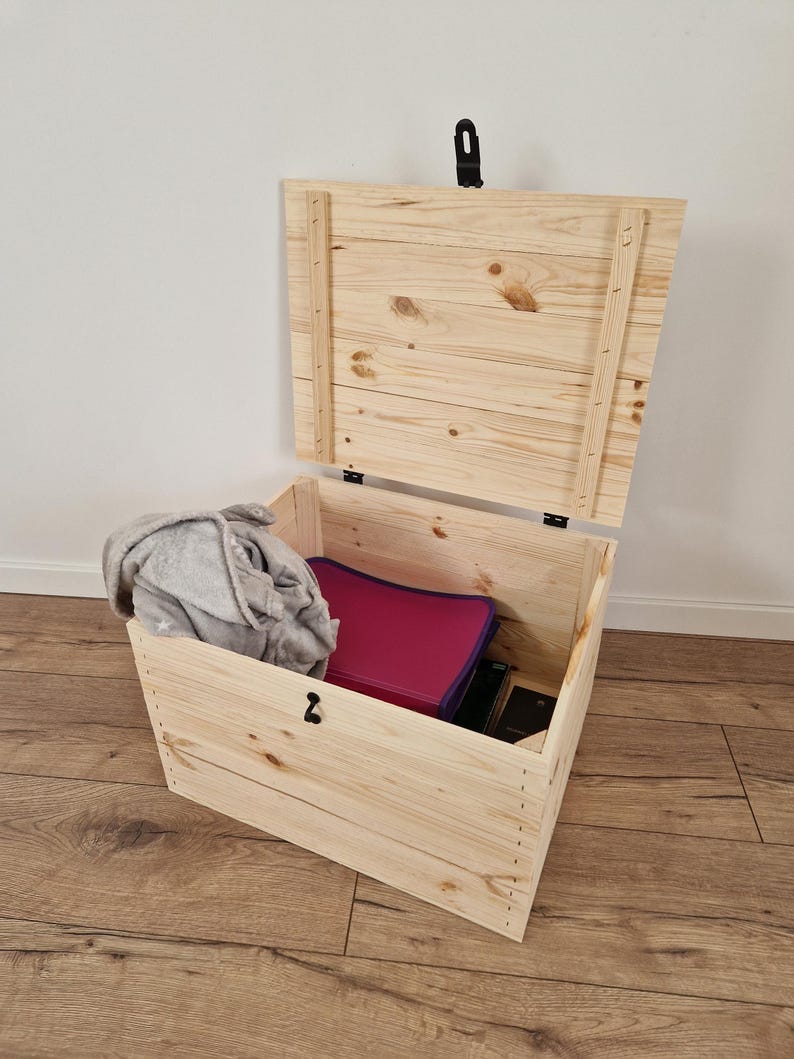 K&ouml;nnte beinhalten: Eine offene, unfertige Holzkiste mit Scharnierdeckel. Im Inneren sind eine graue Decke und ein magenta Ordner zu sehen. Die Kiste ist aus hellem Holz gefertigt und hat einen schwarzen Verschluss. Die Kiste steht auf einem Holzboden.