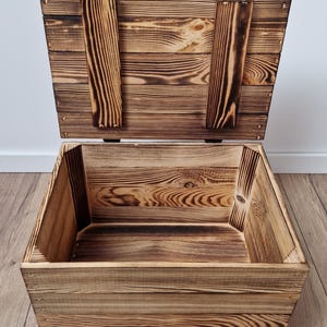 May include: An open wooden storage chest with a hinged lid. The chest is made of light brown wood with darker brown accents and visible wood grain. The lid is propped open, revealing the interior of the box. The chest is sitting on a wooden floor.