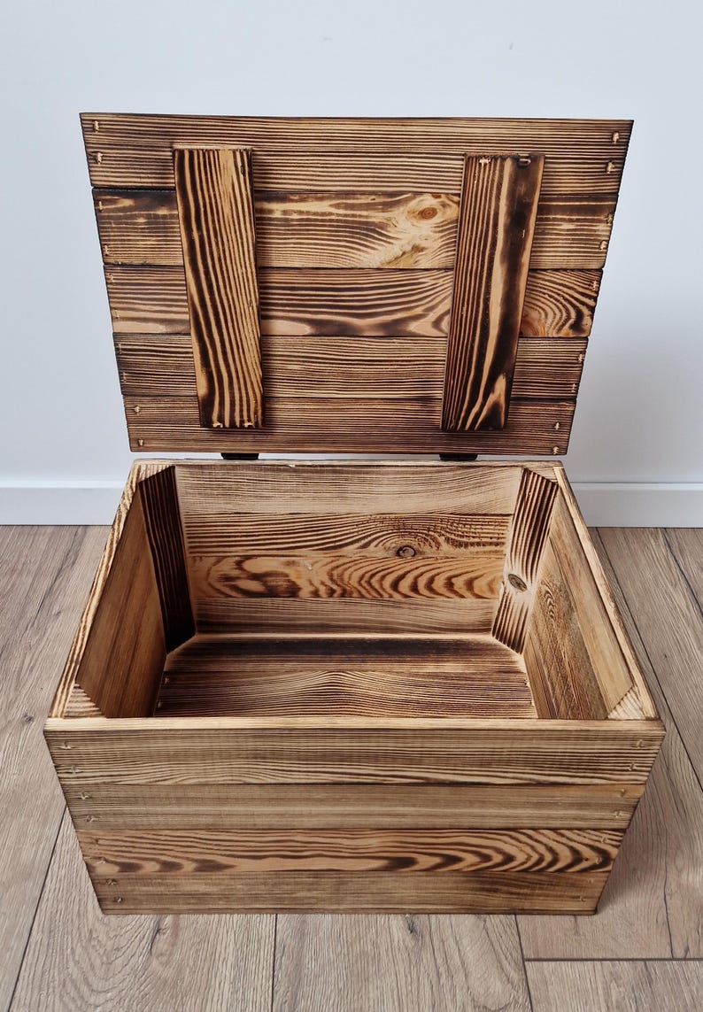 May include: An open wooden storage chest with a hinged lid. The chest is made of light brown wood with darker brown accents and visible wood grain. The lid is propped open, revealing the interior of the box. The chest is sitting on a wooden floor.