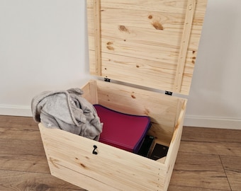 May include: An open, unfinished wooden storage chest with a hinged lid. Inside, a gray blanket, a magenta binder, and other dark items are visible. The chest is made of light-colored wood and sits on a wooden floor.