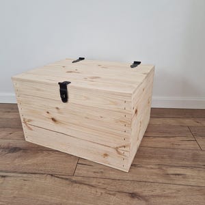 K&ouml;nnte beinhalten: Eine hellfarbene Holzkiste mit Scharnierdeckel und schwarzer Metallbeschl&auml;gen. Die Kiste ist rechteckig und scheint aus unbehandeltem Holz zu bestehen. Die Kiste steht auf einem Holzboden.