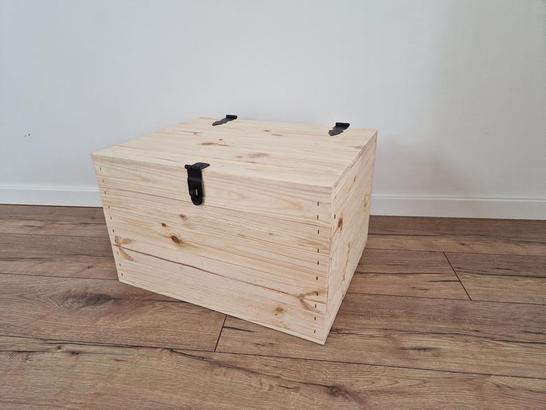 K&ouml;nnte beinhalten: Eine hellfarbene Holzkiste mit Scharnierdeckel und schwarzer Metallbeschl&auml;gen. Die Kiste ist rechteckig und scheint aus unbehandeltem Holz zu bestehen. Die Kiste steht auf einem Holzboden.