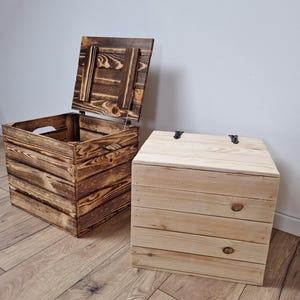 May include: Two wooden storage boxes. One box is open, revealing the interior, and has a dark, burnt wood finish. The other box is closed with a natural wood finish. Both boxes are square-shaped with a slatted design and metal hinges.