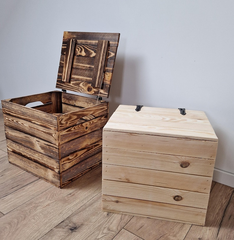 May include: Two wooden storage boxes. One box is open, revealing the interior, and has a dark, burnt wood finish. The other box is closed with a natural wood finish. Both boxes are square-shaped with a slatted design and metal hinges.