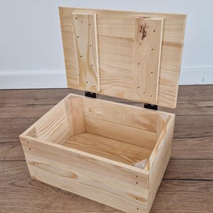 May include: An open, unfinished wooden storage box with a hinged lid. The box is rectangular, constructed from light-colored wood planks, and features visible nail heads. The lid is partially raised, revealing the interior.