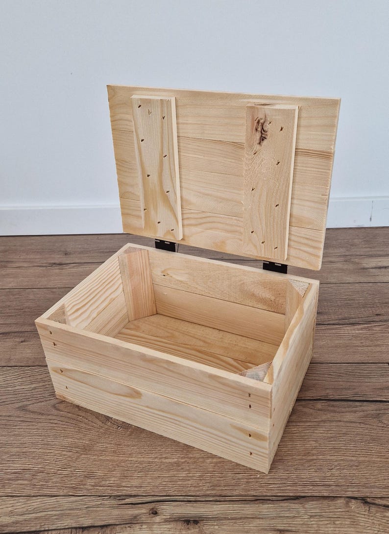 May include: An open, unfinished wooden storage box with a hinged lid. The box is rectangular, constructed from light-colored wood planks, and features visible nail heads. The lid is partially raised, revealing the interior.