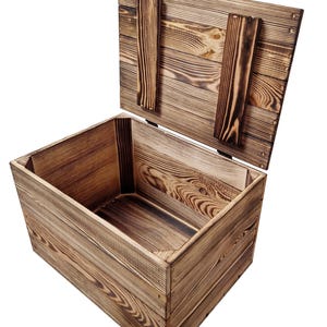 May include: A rustic wooden storage box with a hinged lid, open to reveal the interior. The box is constructed from light brown wood with visible grain and darker, charred accents. The lid is propped open with two wooden supports.