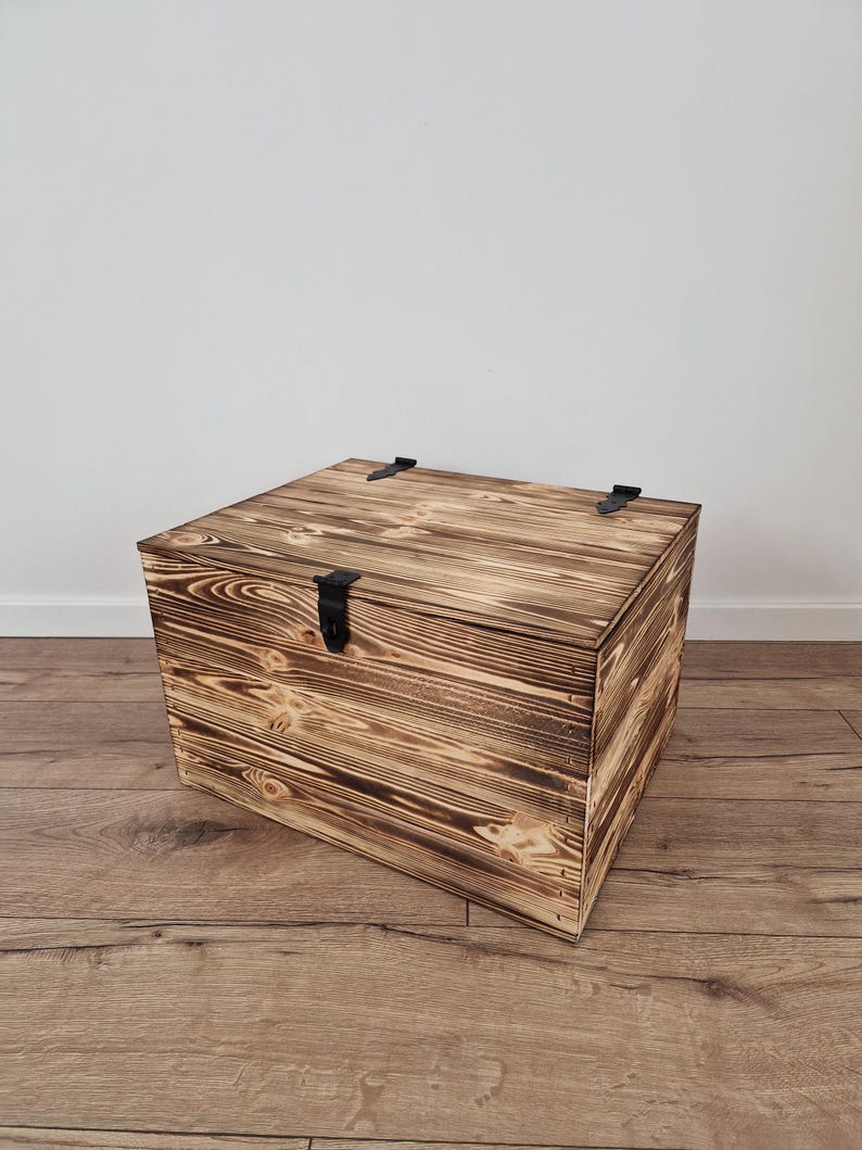 May include: A square wooden chest with a hinged lid and black metal hardware. The chest has a rustic, burnt wood finish, showcasing the natural wood grain. The chest is sitting on a wooden floor.