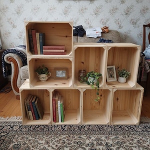 Puede incluir: Un conjunto de cajas de madera apiladas, funcionando como estanterías. Algunas cajas contienen libros, pequeñas plantas en macetas y marcos de fotos. Las cajas de madera clara están sobre una alfombra estampada.