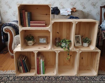 May include: A collection of wooden crates arranged in a stacked configuration, serving as shelves. Some crates contain books, small potted plants, and framed pictures. The crates are made of light-colored wood and are placed on a patterned rug.
