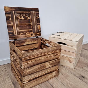 May include: Two wooden storage chests. One chest is open, revealing the interior, and has a dark, charred finish. The other chest is closed and has a lighter, natural wood finish. Both chests are rectangular and appear to be handmade.