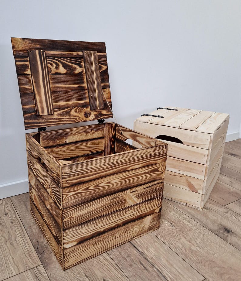 May include: Two wooden storage chests. One chest is open, revealing the interior, and has a dark, charred finish. The other chest is closed and has a lighter, natural wood finish. Both chests are rectangular and appear to be handmade.