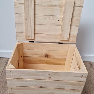 May include: An open, unfinished wooden chest with a hinged lid. The chest is constructed from light-colored planks, with the lid secured by two vertical wooden supports. The interior of the chest is visible, revealing the natural wood grain.