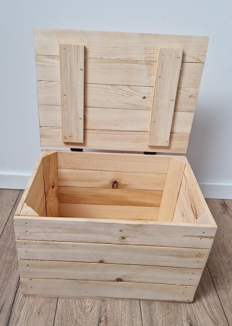 May include: An open, unfinished wooden chest with a hinged lid. The chest is constructed from light-colored planks, with the lid secured by two vertical wooden supports. The interior of the chest is visible, revealing the natural wood grain.