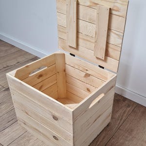 May include: A light-colored wooden storage chest with a hinged lid, open to reveal the interior. The chest has cut-out handles on the sides. The lid is propped open, showing the interior. The chest is made of light-colored wood.