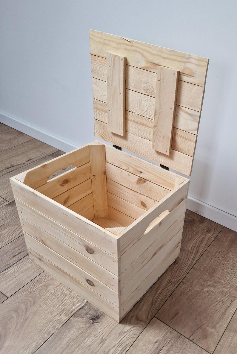 May include: A light-colored wooden storage chest with a hinged lid, open to reveal the interior. The chest has cut-out handles on the sides. The lid is propped open, showing the interior. The chest is made of light-colored wood.