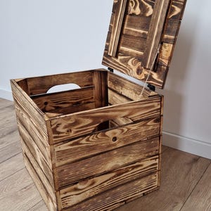 May include: A rustic wooden storage crate with a hinged lid, partially open. The crate is constructed from stained wooden planks, showcasing a natural wood grain pattern. The design includes cut-out handles on the sides.
