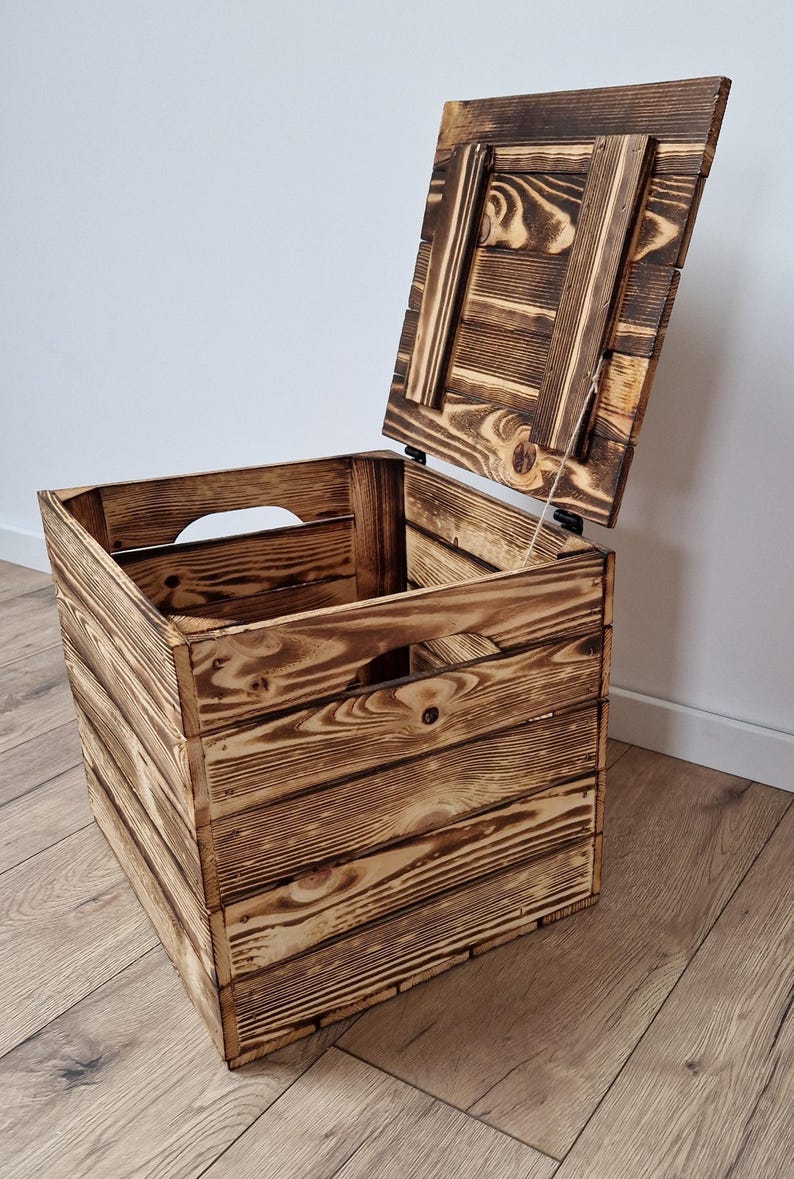 May include: A rustic wooden storage crate with a hinged lid, partially open. The crate is constructed from stained wooden planks, showcasing a natural wood grain pattern. The design includes cut-out handles on the sides.