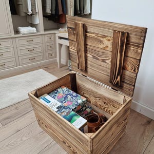 Può includere: Baule portaoggetti in legno aperto con coperchio incernierato, che mostra l'interno pieno di oggetti. Il baule ha una finitura rustica in legno bruciato. All'interno ci sono libri, una scatola a motivi floreali e scarpe di cuoio marrone.