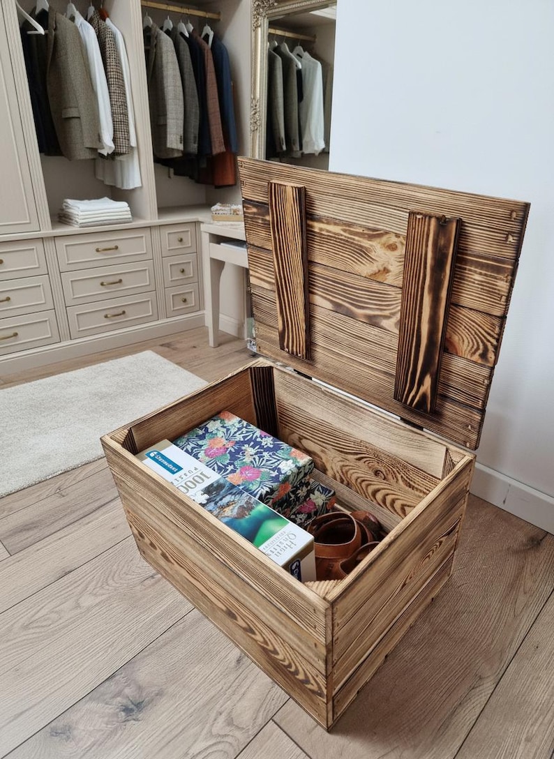 May include: Open wooden storage chest with a hinged lid, showing the interior filled with items. The chest has a rustic, burnt wood finish. Inside are books, a floral patterned box, and brown leather shoes.