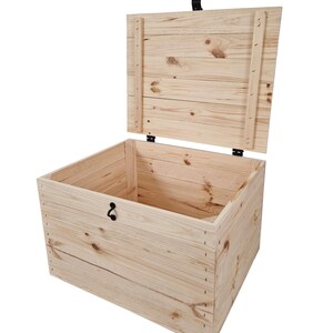 K&ouml;nnte beinhalten: Eine helle Holzkiste mit offenem Deckel. Die Kiste besteht aus Holzbrettern mit sichtbaren Knoten und Maserungen. Sie hat einen schwarzen Riegel und einen Griff. Der Deckel ist leicht ge&ouml;ffnet und gibt den Blick auf das leere Innere frei.
