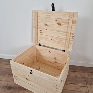 K&ouml;nnte beinhalten: Eine hellfarbene Holz-Truhe mit offenem Deckel. Die Truhe ist aus Holzplanken gefertigt und verf&uuml;gt &uuml;ber einen schwarzen Metallverschluss und einen Griff. Das Innere der Truhe ist sichtbar, und die Holzmaserung ist deutlich zu erkennen.