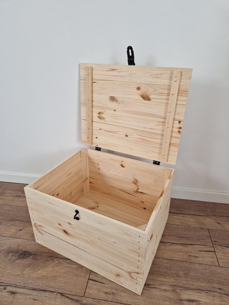 K&ouml;nnte beinhalten: Eine hellfarbene Holz-Truhe mit offenem Deckel. Die Truhe ist aus Holzplanken gefertigt und verf&uuml;gt &uuml;ber einen schwarzen Metallverschluss und einen Griff. Das Innere der Truhe ist sichtbar, und die Holzmaserung ist deutlich zu erkennen.