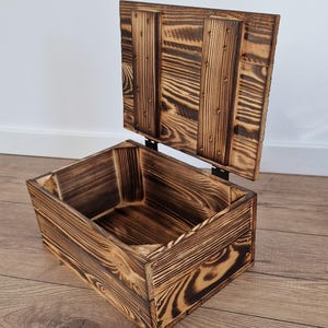 May include: A rustic wooden storage box with a hinged lid, crafted from burnt-look wood. The box is rectangular with visible wood grain and metal hinges. The lid is open, revealing the interior.