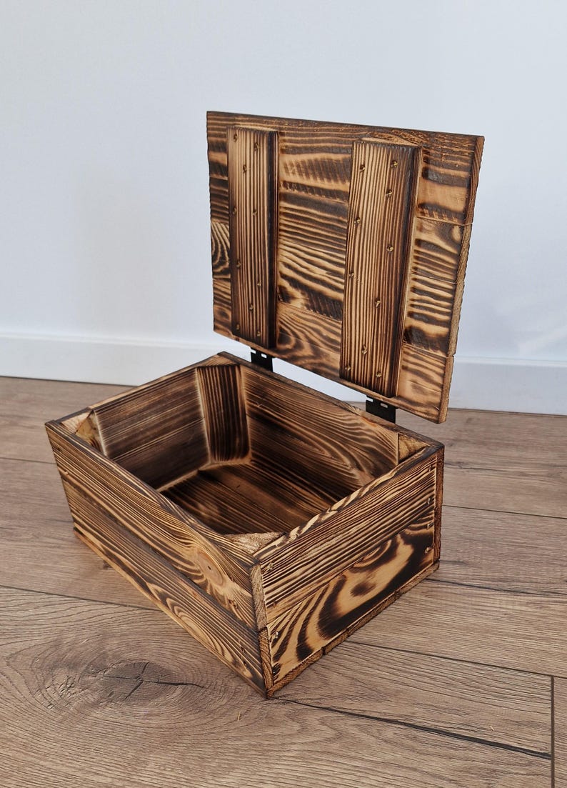May include: A rustic wooden storage box with a hinged lid, crafted from burnt-look wood. The box is rectangular with visible wood grain and metal hinges. The lid is open, revealing the interior.